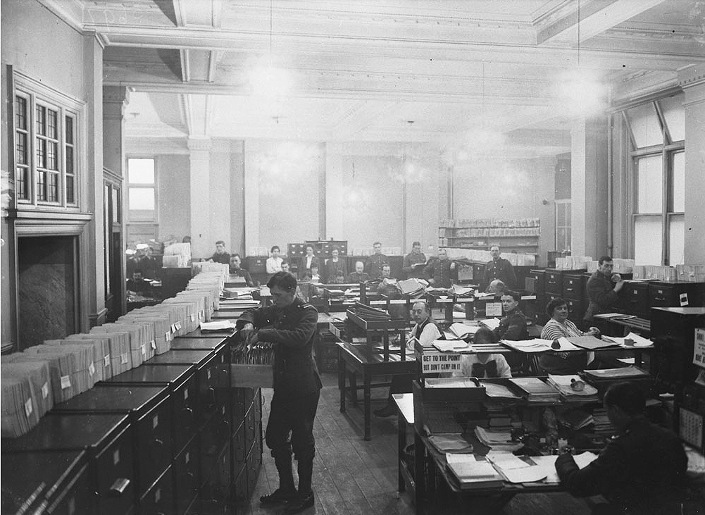 Interior of a large office with a uniformed soldier working at a bank of filing cabinets. Other workers sit at desks.